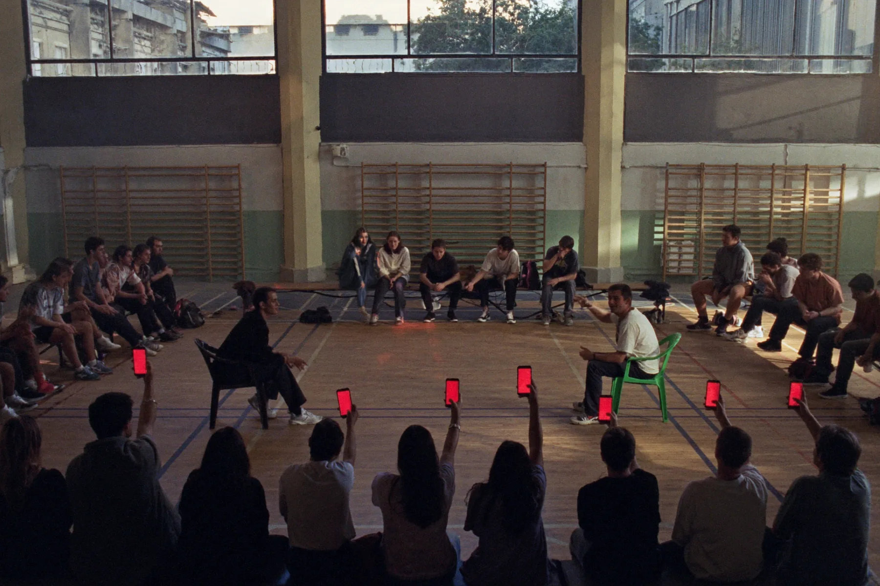A gym seen from above: a circle of teenagers, two chairs facing each other in the middle, and a few raised phones lighting up red