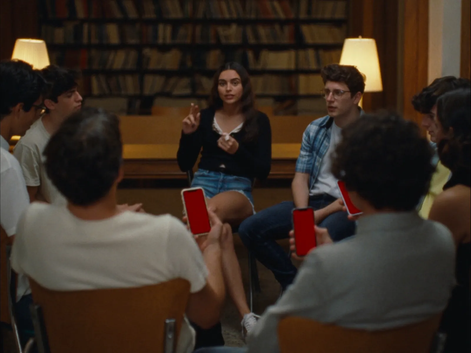 Evening in the library: a circle of students, a girl speaking, some holding up phones with a red screen